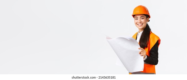 Pleased smiling female asian construction manager following blueprints during building house, looking satisfied at camera, studying architect plan, standing white background in safety helmet