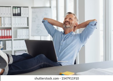 Pleased successful businessman daydreaming or planning his business strategy as he relaxes with his feet up on the desk tilted back in his chair