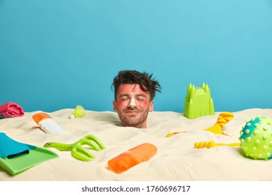 Pleased Sunburnt Man With Protective Sun Cream On Nose, Has Rest At Sandy Beach, Relaxes In Hot Sunny Weather, Surrounded With Different Objects, Has Head Stuck In Sand. Summer Time Concept.