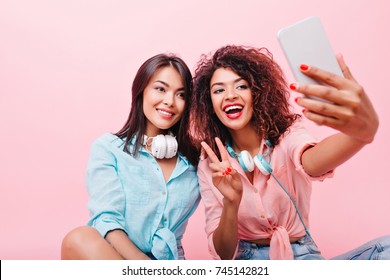 Pleased Tanned Asian Girl Gently Smiling While Her African Friend Making Selfie. Indoor Portrait Of Glad Black Woman With Smartphone Taking Picture Of Herself On Pink Background Near Hispanic Lady.