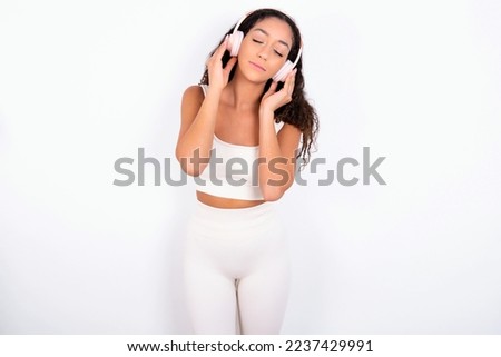 Pleased teen girl with curly hair wearing white sport set over white background enjoys listening pleasant melody keeps hands on stereo headphones closes eyes. Spending free time with music
