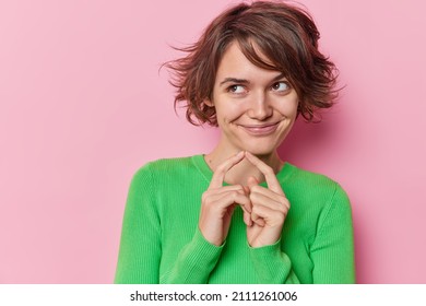 Pleased thoughtful beautiful European woman with short hairstyle keeps index fingers together and schemes something something wears green jumper isolated over pink background has tricky plan