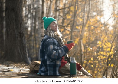 Pleased thoughtful woman rest alone walking in fall forest, drinking hot tea from thermos sitting on wooden path. Satisfied relaxed blonde female in warm clothes enjoys stroll in autumn woodland park - Powered by Shutterstock