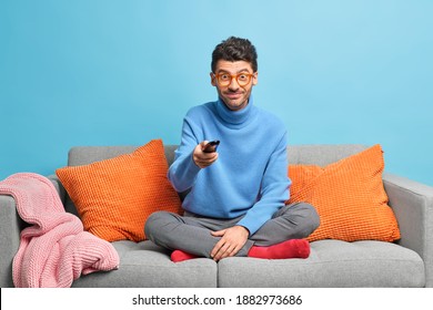 Pleased unshaven man changes TV channels with remote control watches funny program dressed in casual clothes sits crosses legs on comfortable sofa isolated over blue background. Pastime concept