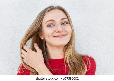 Pleased Woman With Blonde Hair, Luminous Eyes And Gentle Smile Having Dimples On Cheeks Touching Her Beautiful Hair With Hand Dressed In Red Clothes Having Good Mood While Posing Against White Wall