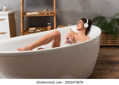 pleased woman in headphones listening music and holding glass of champagne while taking bath - Powered by Shutterstock