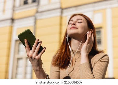 Pleased Woman Listening Music In Wired Earphones And Holding Smartphone Outside