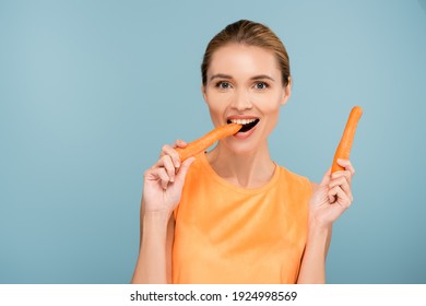 Pleased Woman Looking At Camera While Eating Whole Carrot Isolated On Blue