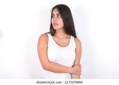 Pleased Young Beautiful Brunette Woman Wearing White Top Over White Wall Keeps Hands Crossed Over Chest Looks Happily Aside