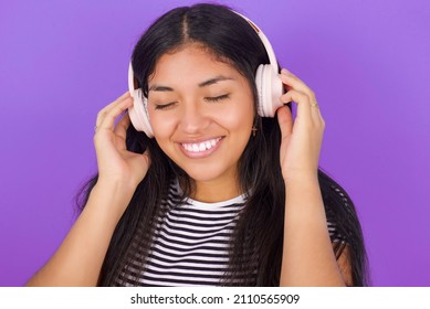 Pleased Young beautiful woman wearing striped T-shirt against purple background enjoys listening pleasant melody keeps hands on stereo headphones closes eyes. Spending free time with music