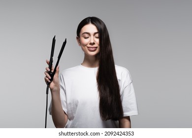 Pleased Young Brunette Woman Holding Hair Straightener Isolated On Grey