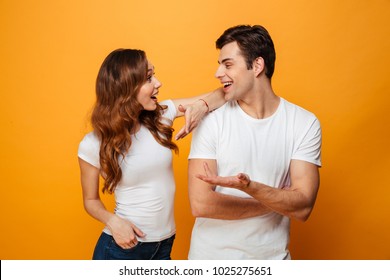 Pleased young lovely couple standing together while talking and looking to each other over yellow background