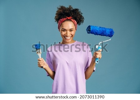 Pleased young mixed race woman enjoying renovation time at home while holding brush and roller. Repair and remodeling concept 