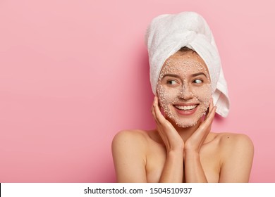 Pleased young woman with delighted expression, applies natural beauty product on face, uncloges pores, has charming smile, looks aside, has wrapped towel on head, has naked body, healthy skin