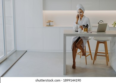 Pleased Young Woman Dressed In Bathrobe Poses Bare Feet At Chair In Spacious Kitchen Sits At Table With Laptop And Cosmetic Products Applies Beauty Patches Under Eyes Talks Via Modern Cellular