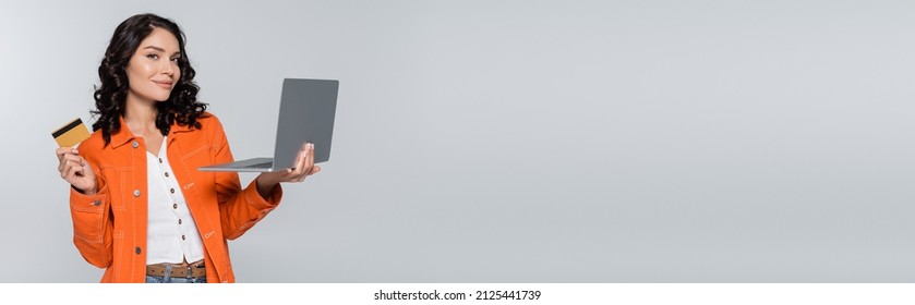 Pleased Young Woman In Orange Jacket Holding Laptop And Credit Card Isolated On Grey, Banner