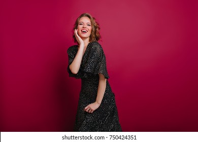 Pleased Young Woman In Vintage Black Dress Posing With Excitement On Claret Background. Indoor Portrait Of Romantic Female Model With Curly Light-brown Hair Smiling During Photoshoot.