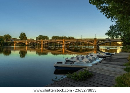 Pleasure boats and pedal boats docked at the wooden plank pier of the Tormes River with the Enrique Esteban Iron Bridge reflected in the water and illuminated beautifully by the golden light of sunset