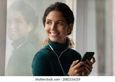 Pleasure to dream. Happy young latin female standing by window in earphones holding mobile phone dreaming by music song. Charming hispanic woman listen to audio book on cell look aside with smile. - Shutterstock ID 2082214219