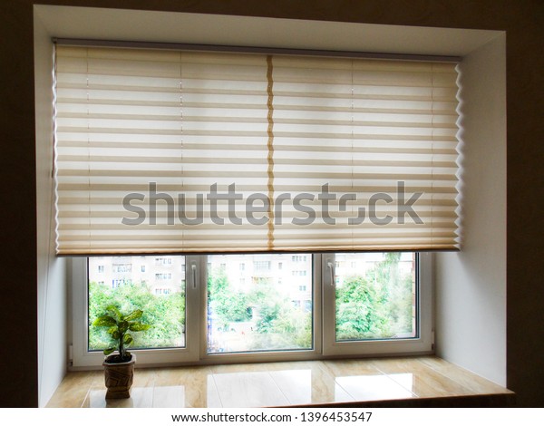 Pleated blinds XL, beige color, with 50mm fold closeup\
in the window opening in the interior. Home blinds - modern bottom\
up privacy shades half raised on apartment windows.          \
