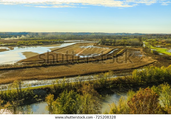 Plowed field in the fall; water appears on\
the left; blue sky above the field; dust raised by a driving car in\
the background on the left; aerial\
view