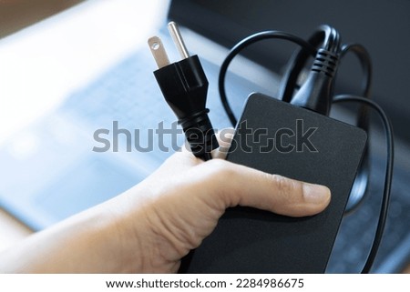 Plug in power outlet adapter cord charger on a woman's hand, with blurred behide of the laptop computer on wooden floor.