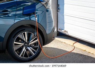 A plug-in hybrid electric car (PHEV), parked in front of the home garage door, is plugged in with a charging cable to recharge the battery.