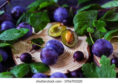a plum cut in half lies on a piece of wood. There are whole plums lying around with green leaves on the branches. harvesting the crop