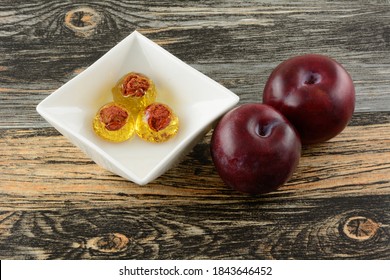 Plum Hard Candy Drops With Plum Pit In Center In White Candy Dish And Fresh Raw Plum Fruit On Table