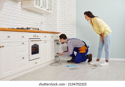 Plumber crouching on the floor in a modern white kitchen and using a drain cable to clean a clogged sink pipe. Young girl calls the plumber to fix the problem of a clogged drain under her kitchen sink