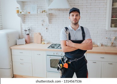 Plumber In Kitchen. Professional Look On Camera And Pose. Hands Crossed. Tools On Belt Aside.