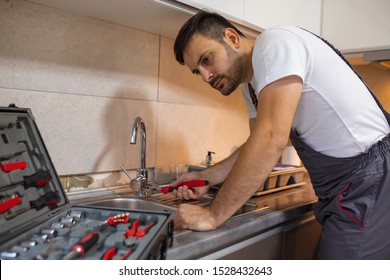 Plumber Man Fixing Kitchen Sink. Handsome Plumber At The Kitchen. Reparation. Handsome Plumber Repairing Sink Pipes In Kitchen. Serious Male Plumber In Working Overall Fixing Sink In Kitchen