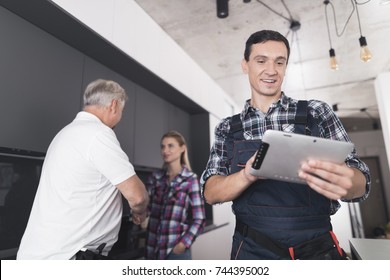 The plumber is posing in the kitchen. He looks at the tablet and smiles. Behind him is a client and communicates with the second plumber. A black toolbox is next to it. - Powered by Shutterstock - Get 15% off with code: PIKWIZARD15