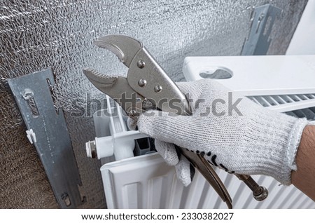 Plumber with a wrench repairing a heating radiator. Foil thermal insulation on the wall behind the heating radiator at home. 
