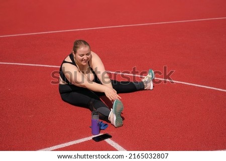 Plump fat woman is doing sports on rubber covering of stadium. The person does stretching before fitness. Outdoor training on summer morning. Striving for weight loss , maintaining healthy lifestyle