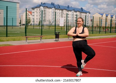 Plump Fatty Chubby Woman Doing Yoga Standing In A Tree Pose At A Sports Outdoor Stadium . Female Keeps Balance By Doing Fitness. Sports Ground With Red Rubber Coating