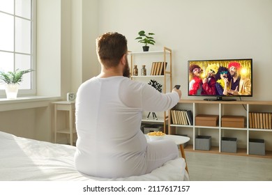 Plump Ginger Guy Wakes Up In Morning And Switches Television With Remote Control. Funny Fat Man In PJs Sitting In Bedroom At Home And Watching Entertainment Channel On Widescreen HD LED TV, Back View