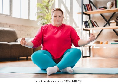 Plump Obese Caucasian Fat Woman Meditating In Sporty Outfit At Home. Fat Woman Sitting And Relaxing In Lotus Position. Chubby Woman Doing Yoga At Home For Losing Weight