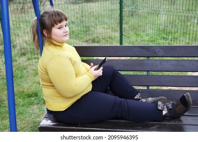 Plump Thoughtful Girl In Yellow Sweater Sits Alone On Bench Of Swing Outdoors, Hold Smartphone, Looks Aside And Thinks