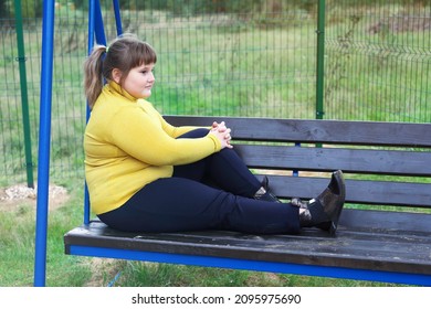 Plump thoughtful girl in yellow sweater sits alone on bench of swing outdoors, looks aside and thinks
