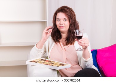 Plump woman at home eating chocolates and drinking wine