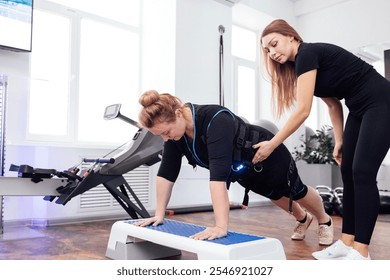 Plump woman middle age in ems suit is engaged on step platform with personal fitness trainer. Blonde woman doing push ups from the stepper. Electrical muscle stimulation workout in modern gym. - Powered by Shutterstock