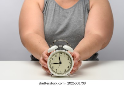 Plump Woman Sits At A Table And Holds A Round White Alarm Clock. The Concept Of Fractional Nutrition, Time To Lose Weight, Adherence To Sleep