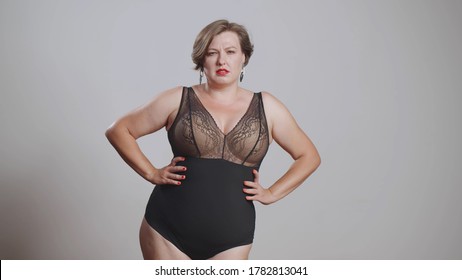 Plump Young Woman In Black Body Stockings Standing On Grey Background With Hands On Hips Looking At Camera. Beautiful Plus Size Female Model In Lingerie With Copy Space.