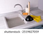 A plunger and yellow rubber gloves on top of a clogged kitchen sink