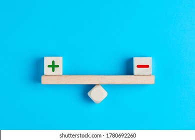 Plus and minus or positive and negative symbols on wooden blocks are in balance on a wooden seesaw. Blue background, flat lay view. Pros and cons equilibrium in decision making under uncertainity. 