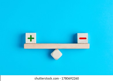 Plus And Minus Or Positive And Negative Symbols On Wooden Blocks Are In Balance On A Wooden Seesaw. Blue Background, Flat Lay View. Pros And Cons Equilibrium In Decision Making Under Uncertainity. 