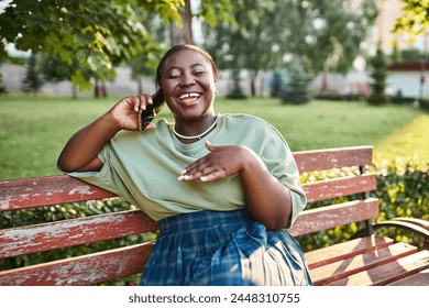 Plus size African American woman in casual attire, sitting on a bench outdoors in the summer, talking on a cell phone. - Powered by Shutterstock