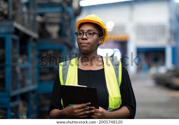 Plus size black female worker wearing safety\
hard hat helmet inspecting old car parts stock while working in\
automobile large warehouse