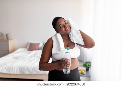 Plus Size Black Woman With Bottle Of Water Wiping Sweat With Towel After Domestic Workout, Indoors. Plump African American Lady Staying Hydrated During Sports Training. Weight Loss, Slimming Concept